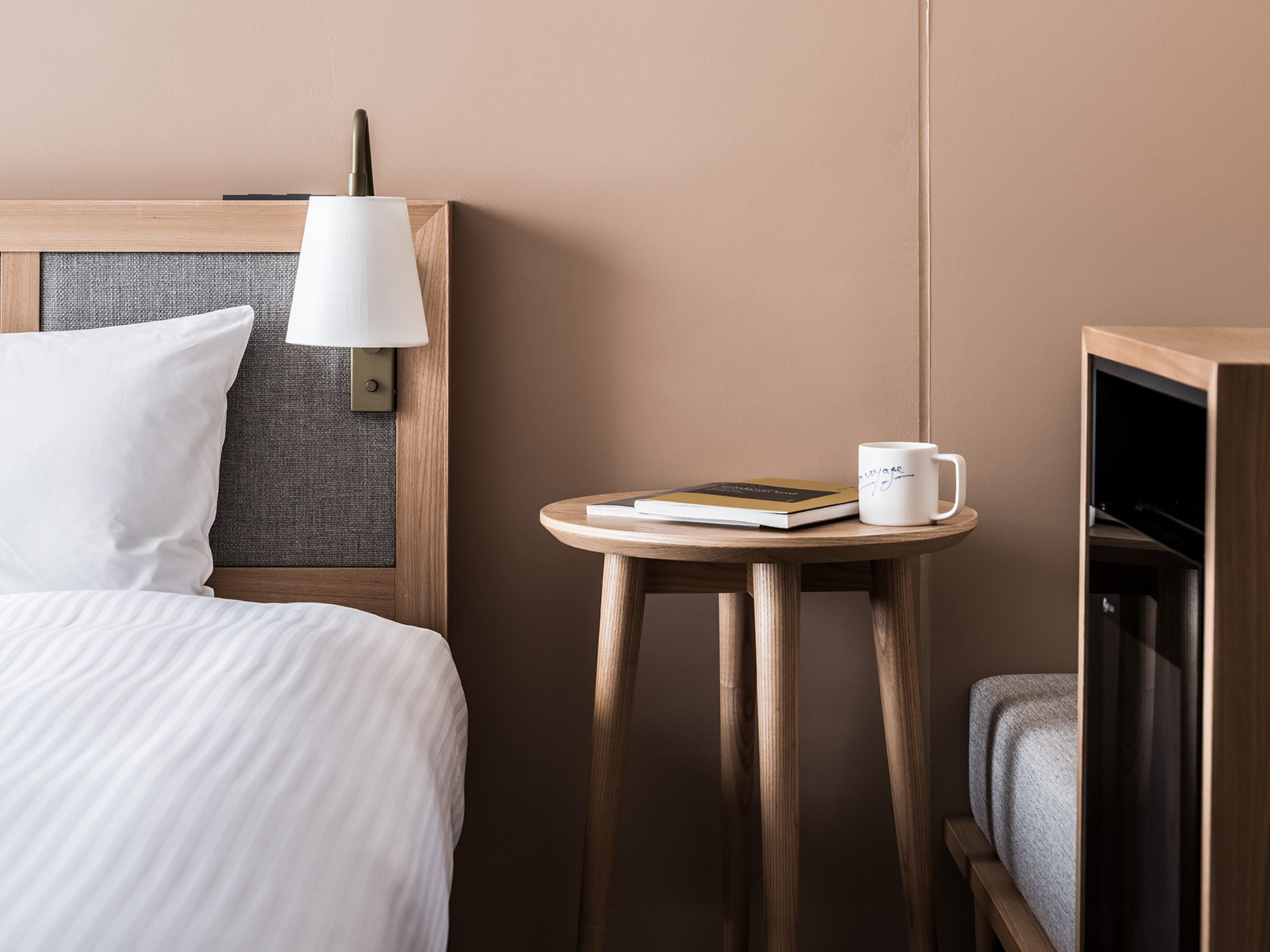 A neatly arranged hotel room with a bed, a bedside lamp, a small table with a book and a mug, and a television.
