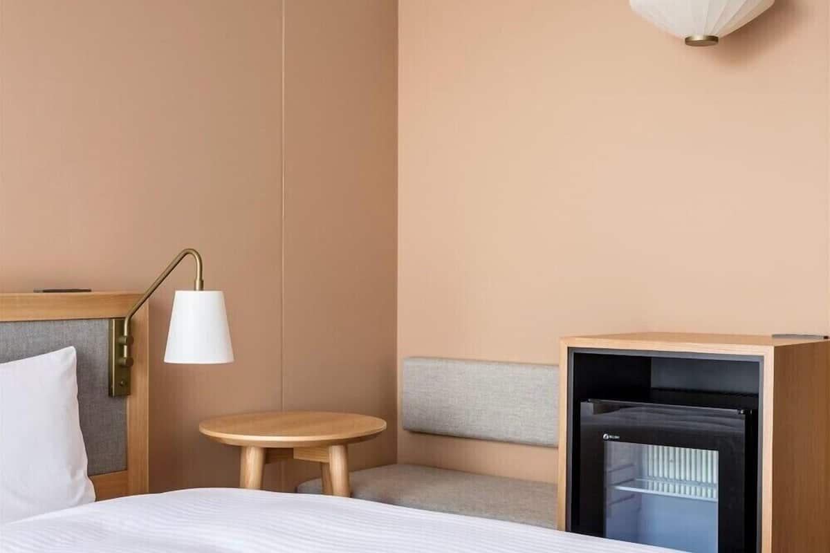 A hotel room with a bed, a bedside table, a lamp, a small round table, a bench, and a minibar.