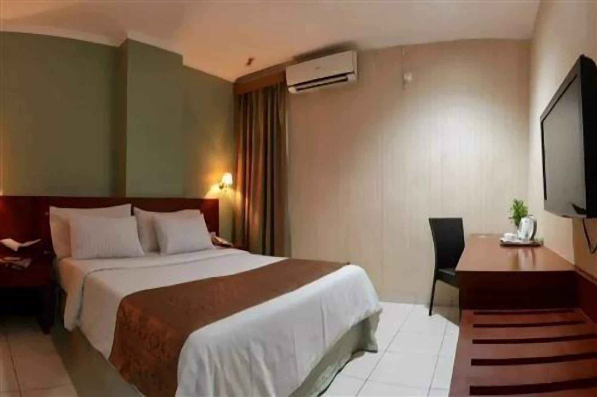 A hotel room with a bed, a desk, a chair, a TV, and an air conditioning unit.