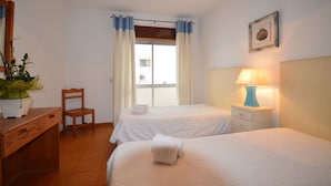Ein Hotelzimmer mit zwei Betten, einem Holztisch, einem Stuhl, einer Topfpflanze, einem Fenster mit Vorhängen, einer Wandlampe und einem gerahmten Bild.