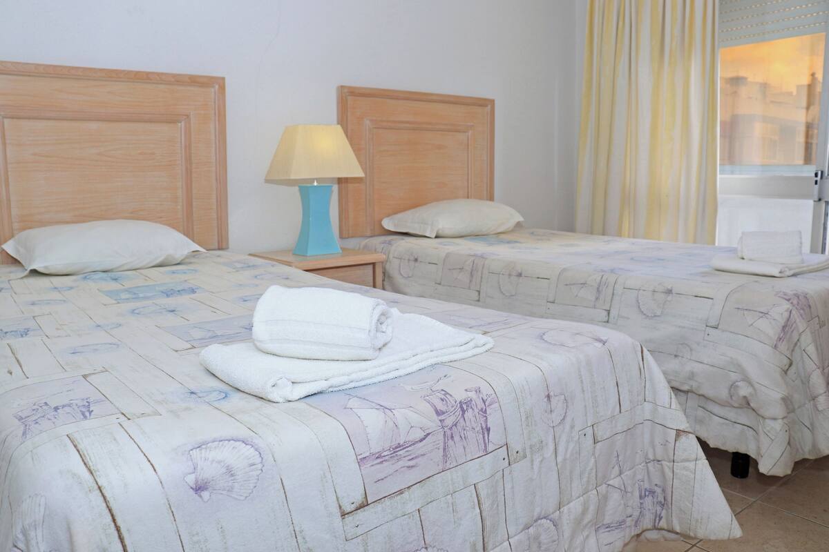 A hotel room with two beds, a nightstand with a lamp, and a window with curtains.