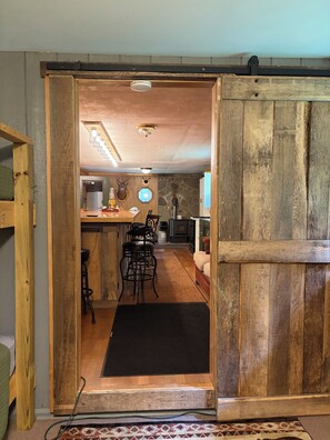 Un interior rústico con una puerta de madera que da a una zona de cocina con una barra, taburetes y una pared de piedra.