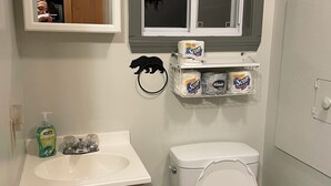 Une salle de bain avec un lavabo, des toilettes et un miroir, tous de couleur blanche. Du papier toilette et du savon liquide sont fixés au mur.