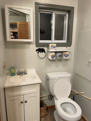 Un baño con lavamanos, inodoro y espejo blancos. Hay papel higiénico y jabón para manos en la pared.