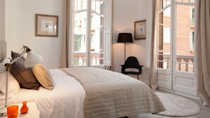 A bedroom with a bed, a desk with a lamp, a chair, and a view of the outside.
