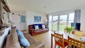 A spacious living room with a brown leather sofa, a wooden dining table with chairs, a television, and a large window with a view of the outdoors.