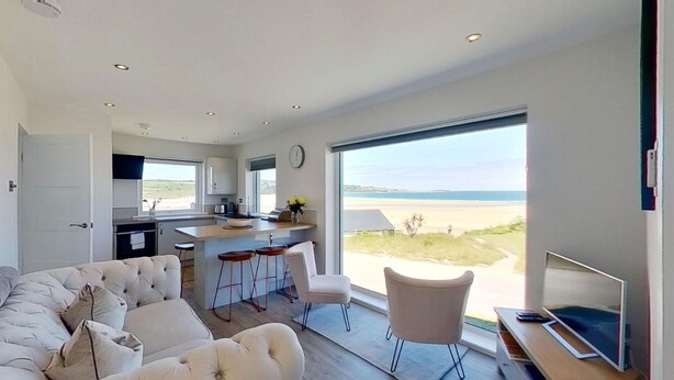 A modern living room with a large window overlooking a beach, a kitchen area, and a seating area with a sofa and bar stools. - B27 The Lair, Riviere Towans (Hayle)