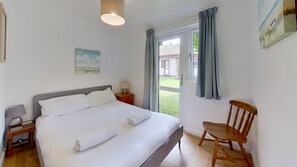 A bedroom with a bed, a chair, a nightstand, a lamp, a painting, and a window with curtains. - 09 Trevithick Court, Tolroy Manor (Hayle)