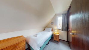 A small, tidy bedroom with a single bed, a wooden dresser, a nightstand with a lamp, and a window with curtains.