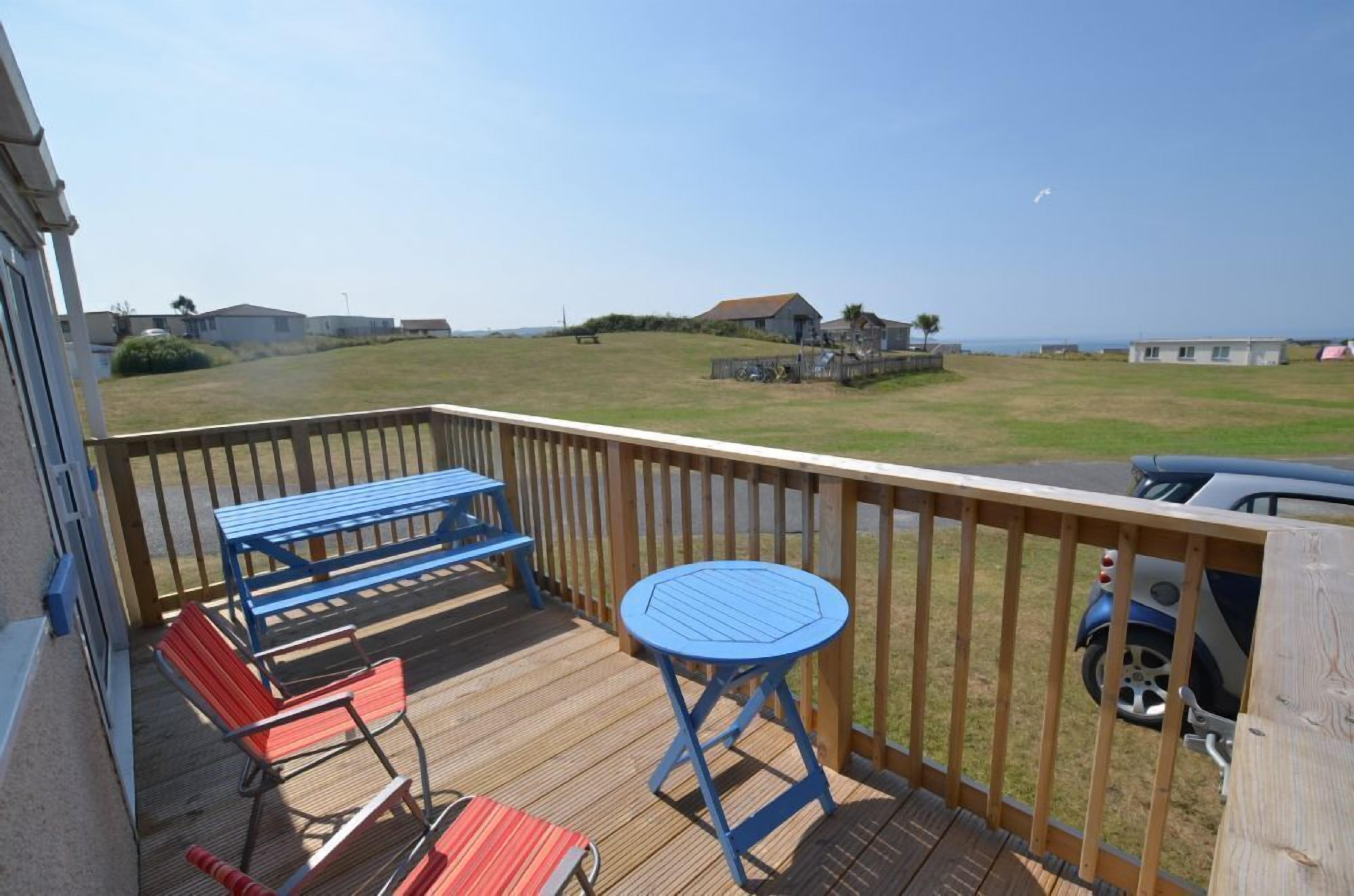 Una terraza de madera con sillas azules y rojas, una mesa pequeña y vista a la playa y casas al fondo.