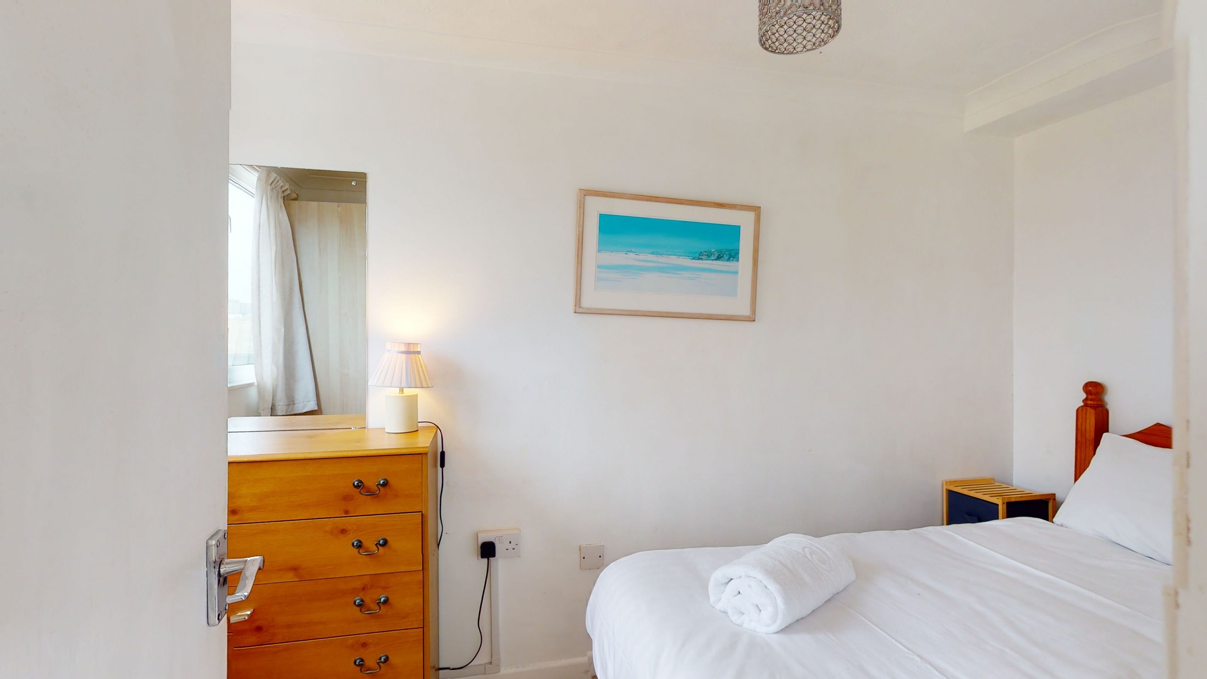 Een slaapkamer met een houten dressoir, een nachtkastje, een bed met een opgerolde handdoek, een schilderij van een strandgezicht en een hangende lamp.