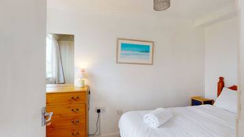 Een slaapkamer met een houten dressoir, een nachtkastje, een bed met een opgerolde handdoek, een schilderij van een strandgezicht en een hangende lamp.