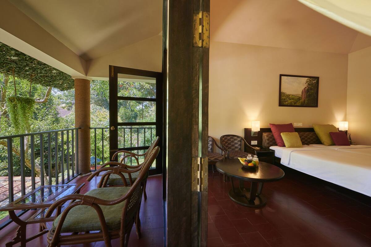 A room with a bed, a sofa, and a dining table. There is a balcony with chairs and a railing.