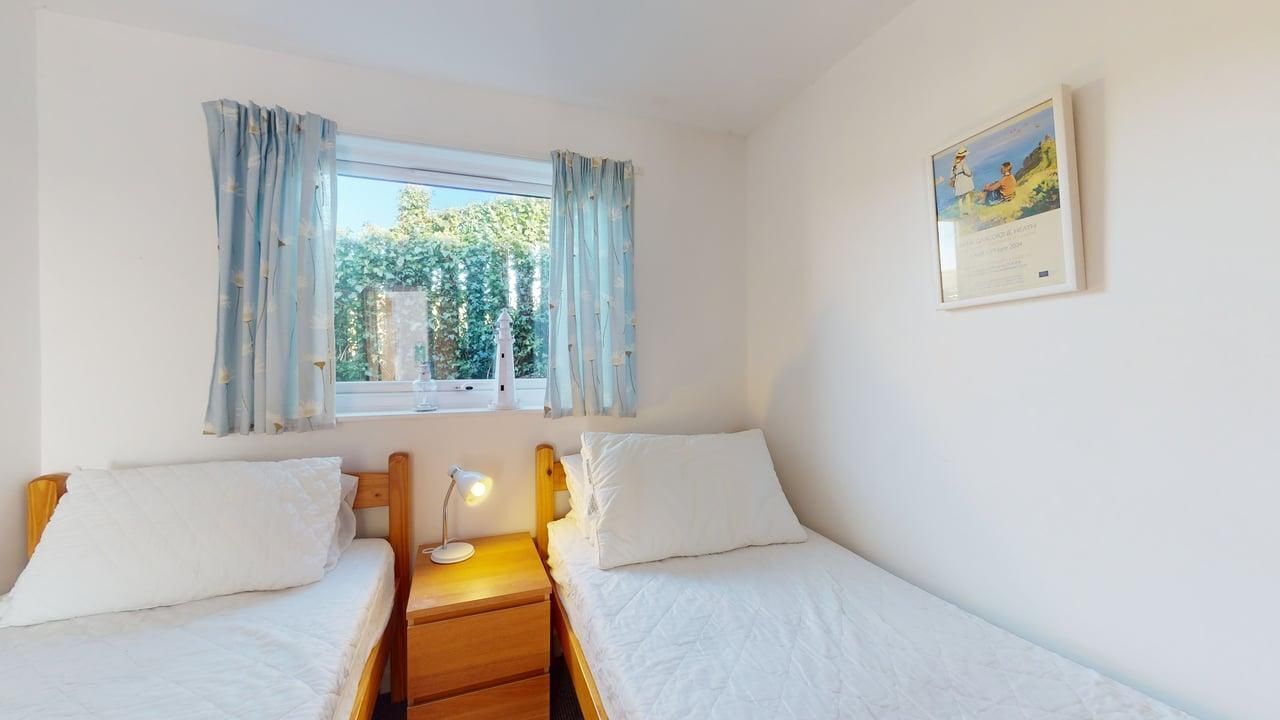 A bedroom with two single beds, a nightstand with a lamp, a window with curtains, and a framed picture on the wall.
