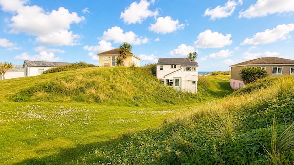 A coastal residential area with houses on a grassy hill, clear skies, and a calm sea in the background.