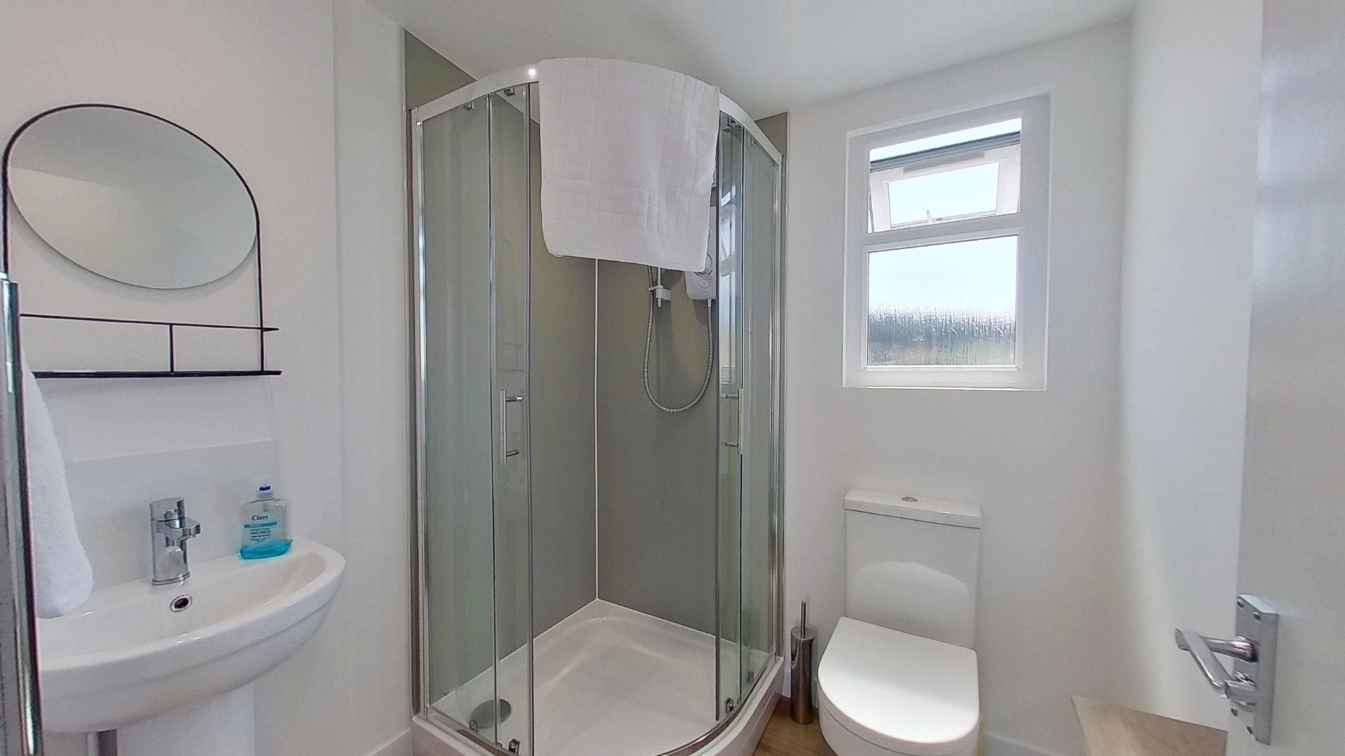 Ein modernes Badezimmer mit einer gläsernen Duschkabine, einem weißen Waschbecken, einer Toilette und einem Fenster, das natürliches Licht hereinlässt.