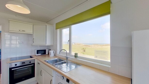 Cucina con mobili bianchi, un forno a microonde, un bollitore e un lavandino. Si vede una finestra con vista sul mare e un tendone verde.