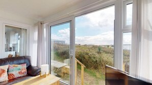 Un salon moderne comprenant un canapé, une table et un téléviseur. Il est doté de grandes portes-fenêtres coulissantes qui donnent sur un espace extérieur offrant une vue sur la campagne.