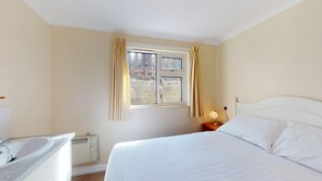 A hotel room with a bed, a nightstand with a lamp, a window with curtains, a radiator, and a bathroom with a sink. - G21 Wellington, Riviere Towans (Hayle)