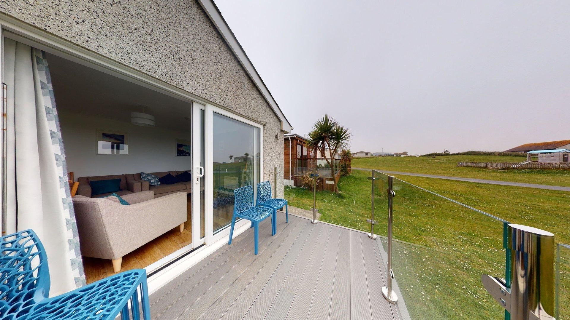 A modern outdoor living area with a glass door leading to a furnished room, a wooden deck, and a view of the countryside.
