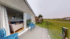 A modern outdoor living area with a glass door leading to a furnished room, a wooden deck, and a view of the countryside.