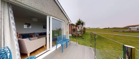 A modern outdoor living area with a glass door leading to a furnished room, a wooden deck, and a view of the countryside.