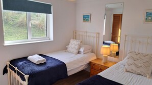 Une chambre avec deux lits simples, une table de chevet avec une lampe, une fenêtre donnant sur un paysage verdoyant et un miroir qui reflète la pièce.