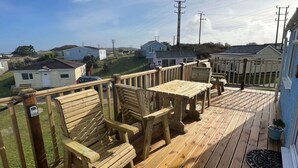 A wooden deck with a table and chairs, overlooking a residential area with houses and power lines. - E08 Boness (E8), Riviere Towans (Hayle)