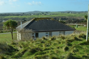 A small house with a sloped roof, surrounded by grassy terrain and a distant view of hills.