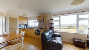 A kitchen with wooden cabinets, a dining table, a black leather sofa, and a window with a view. - D55 Wheal Lucy, Riviere Towans (Hayle)