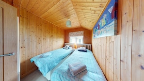 Een kamer met houten wanden en plafond, twee eenpersoonsbedden met blauw beddengoed, een raam met witte gordijnen en een schilderij aan de muur.