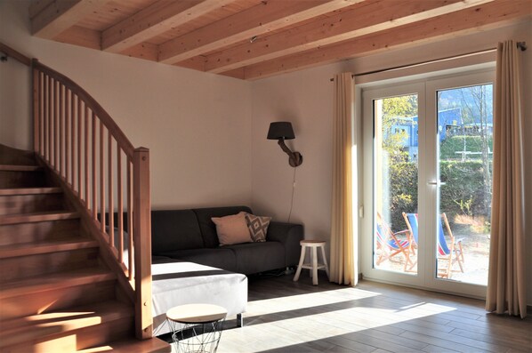 A room with a wooden staircase, a sofa, a small table, and a view through a glass door to an outdoor area with chairs.