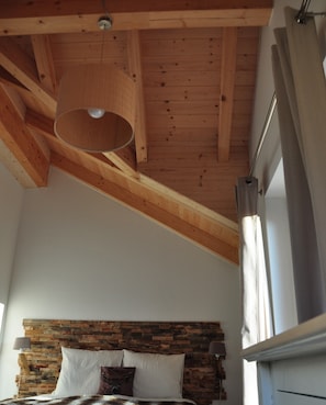 A bedroom with a sloped ceiling, a wooden headboard, a bed with pillows, a hanging light fixture, and a window with curtains.