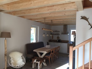 A rustic interior with a wooden ceiling, a dining area with a table and chairs, a sofa, and a kitchen area with cabinets and a hanging light fixture.