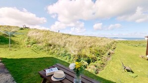 A grassy outdoor area with a wooden bench, a book, a hat, and a vase with flowers. - B38 Delamar, Riviere Towans (Hayle)
