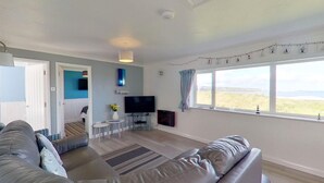 A modern living room with a grey sofa, a flat-screen TV, and a large window with a view of the countryside. - B38 Delamar, Riviere Towans (Hayle)