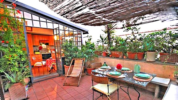 Un patio exterior con una mesa de comedor lista para dos personas, rodeado de macetas con plantas y una pérgola de madera.