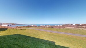 A coastal landscape with a paved path leading to a grassy area, houses in the background, and a clear blue sky. - B04 Beachcroft (B4), Riviere Towans (Hayle)
