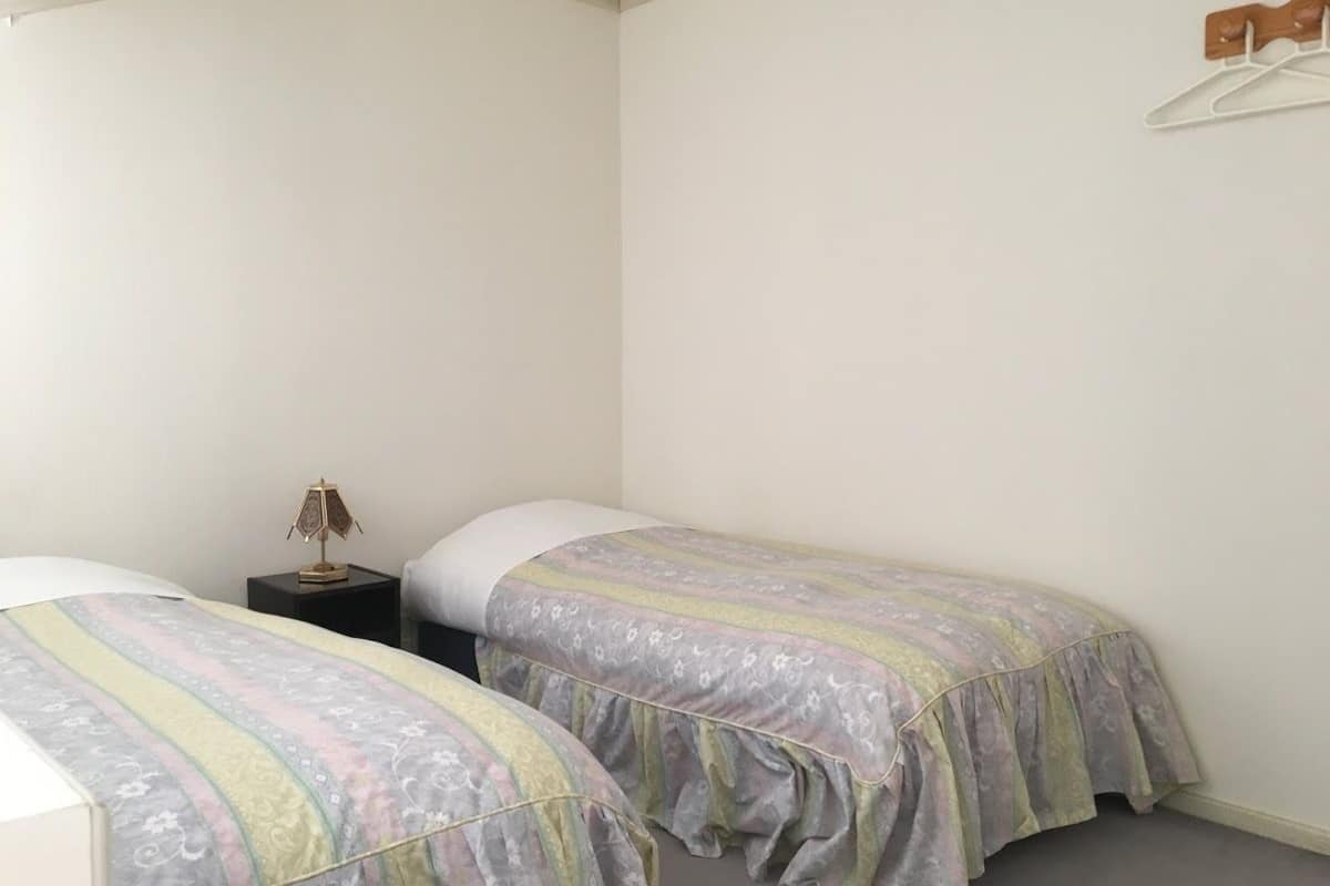 Um quarto pequeno e simples com duas camas de solteiro, uma mesa de cabeceira com abajur e uma parede sem decoração.
