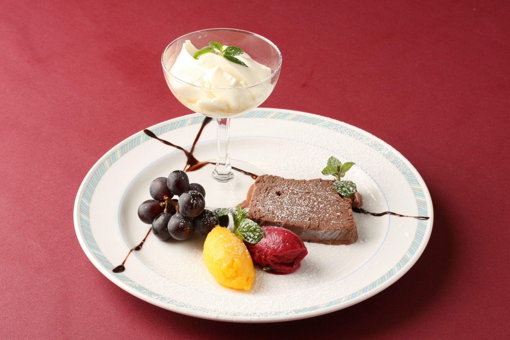 Un plato de postre con mousse de chocolate, uvas, una rodaja de limón y una fresa, servido sobre un plato blanco con borde azul.
