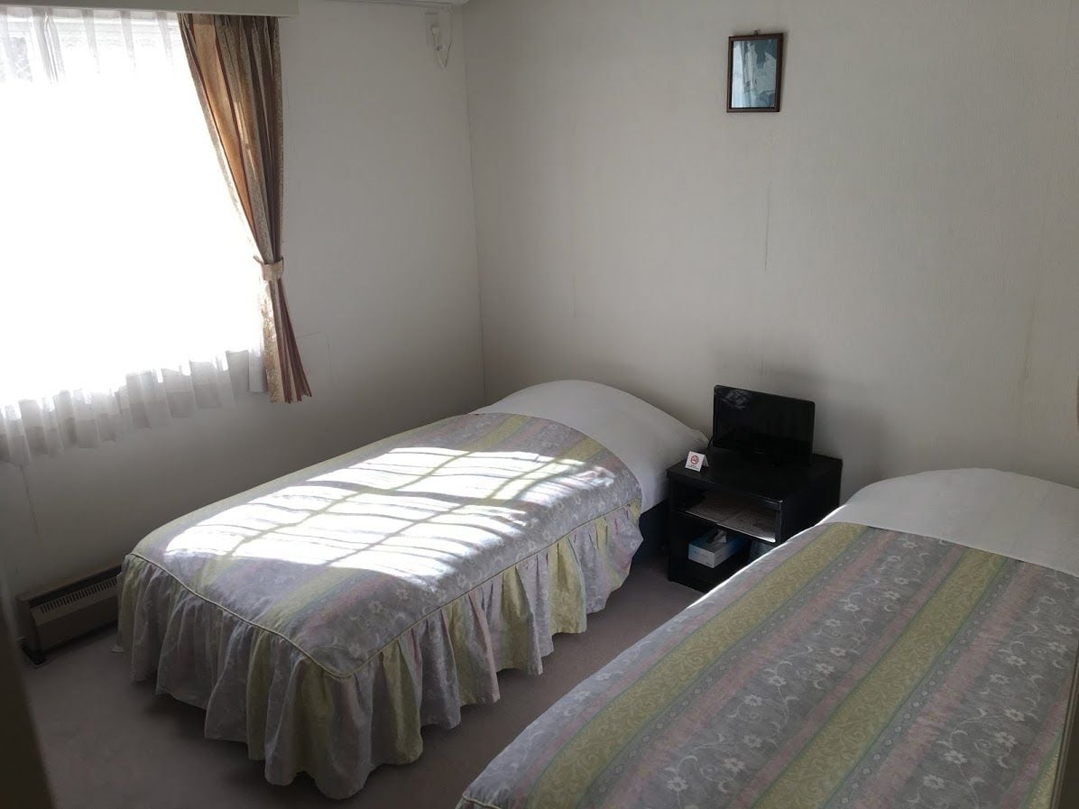 A room with two single beds, a heater, a small desk, and a window with curtains.
