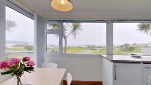 A room with a dining table, chairs, a vase with flowers, and a view of palm trees and houses through large windows. - D29 Westbourne, Riviere Towans (Hayle)