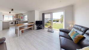 A modern living room with a sofa, a dining table, and a TV. There is a kitchen area with a washing machine and a dining table. The room has a large sliding glass door leading to a garden. - B51 Blue Bay, Riviere Towans (Hayle)