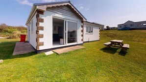 A single-story house with a white exterior and stone accents, a sliding glass door entrance, and a picnic table in the yard. - B51 Blue Bay, Riviere Towans (Hayle)