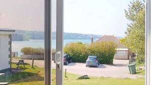 A view through a glass door of a parking area with two cars, a grassy area, and a building in the background. - B51 Blue Bay, Riviere Towans (Hayle)