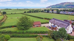 A rural landscape with a stone house featuring a thatched roof, surrounded by green fields and a mountain in the background. - Splatt Barn, Porlock (Porlock)