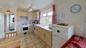 A compact kitchen with a microwave, oven, and sink. There is a clock on the wall and a window with curtains. - A20 Vardo, Riviere Towans (Hayle)