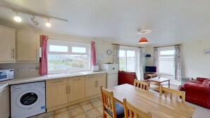 A compact kitchen with a washing machine, a dining area with a table and chairs, and a red sofa. - A20 Vardo, Riviere Towans (Hayle)