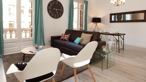 A modern living room with a brown sofa, white chairs, a glass coffee table, a large wall clock, and a dining area with a table set for two. - Stylish apartment in city center 23 (Barcelona)