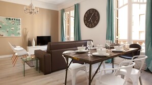 A modern living room with a brown sofa, white chairs, a wooden dining table set for four, and a large wall clock. - Stylish apartment in city center 23 (Barcelona)